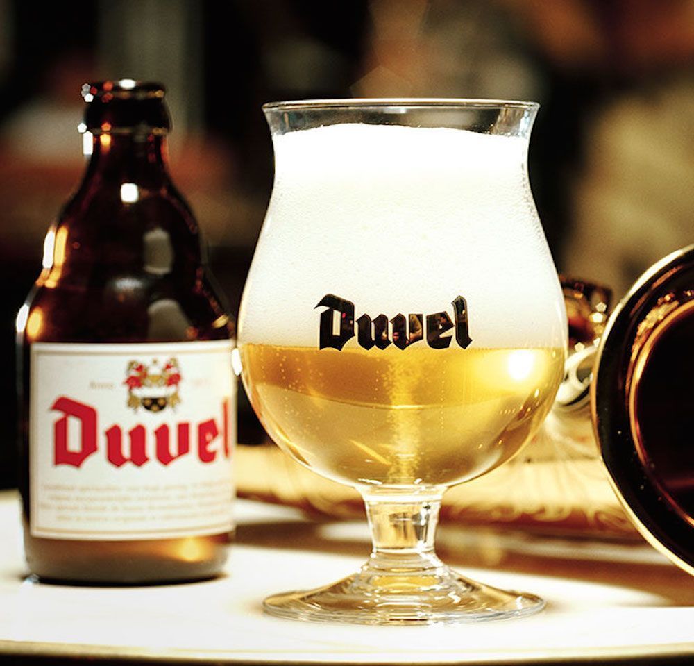 Botella y vaso de cerveza Duvel, líquido dorado con espuma blanca, sobre superficie blanca.