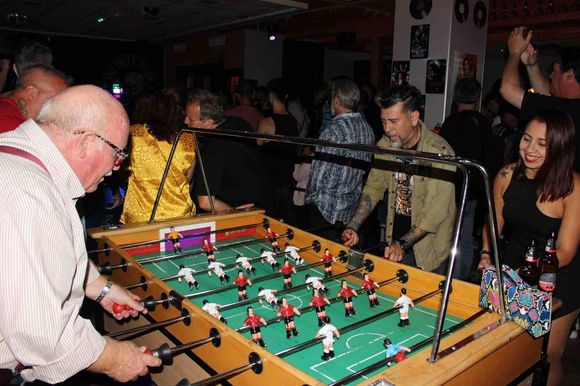 Hombres jugando al billar en un bar. Un hombre de pelo largo está de pie cerca de la mesa, mientras otro prepara un tiro.