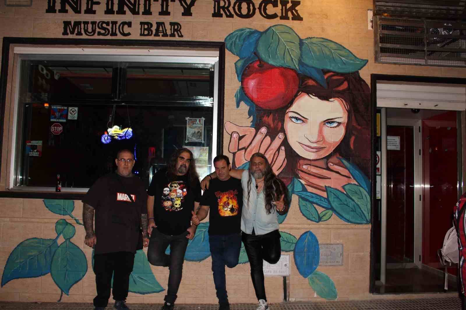 Cuatro personas posan afuera del Infinity Rock Music Bar, frente a un mural de una mujer con frutas y hojas.