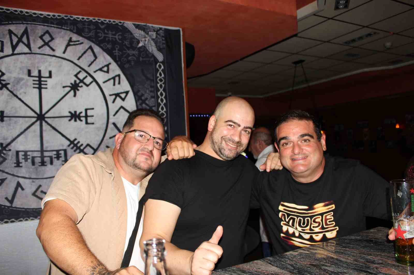 Tres hombres sonrientes, abrazados, cerca de un bar. Un símbolo vikingo está en la pared detrás de ellos.