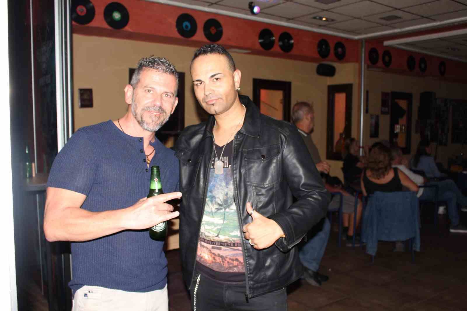 Dos hombres en un bar, uno con una cerveza en la mano, sonriendo. Otro con chaqueta de cuero levanta el pulgar. Paredes decoradas con discos.