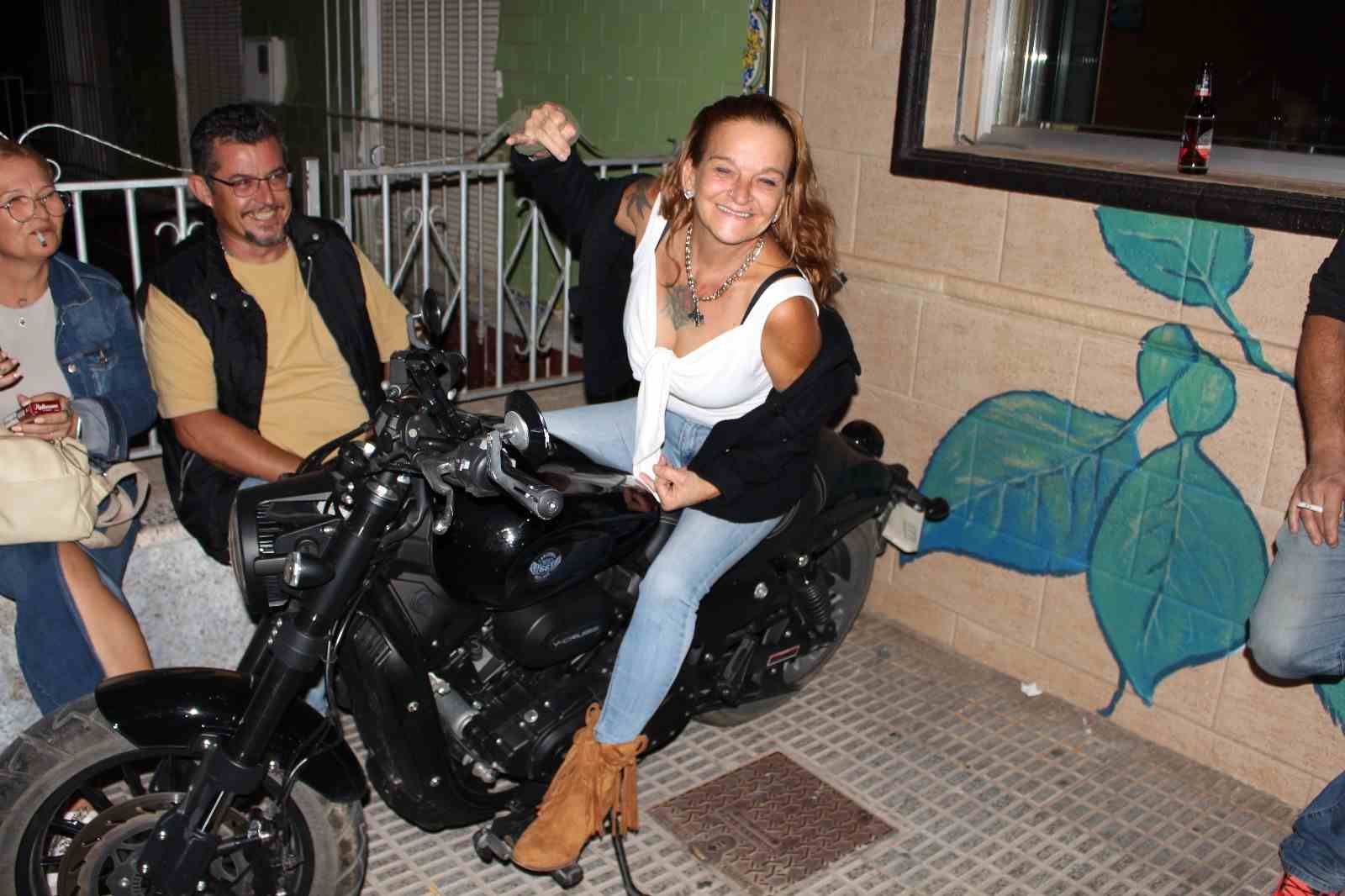 Una mujer posa en una motocicleta negra, levantando el brazo. Dos personas la observan. Bar.