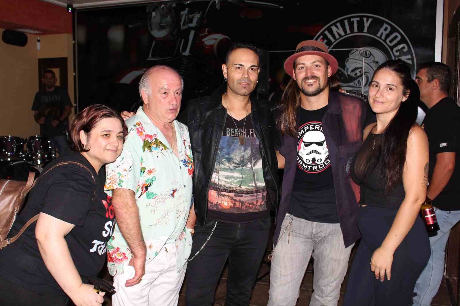 Cinco personas posan para una foto en un local. Dos llevan camisetas de bandas. El local cuenta con bar y batería.