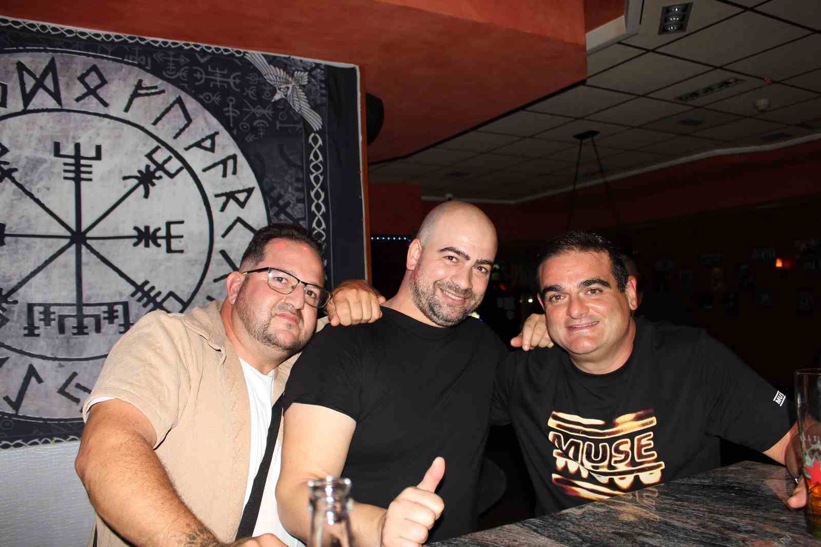 Tres hombres sonriendo, abrazados, en un bar. Detrás de ellos, una pared con un símbolo vikingo.