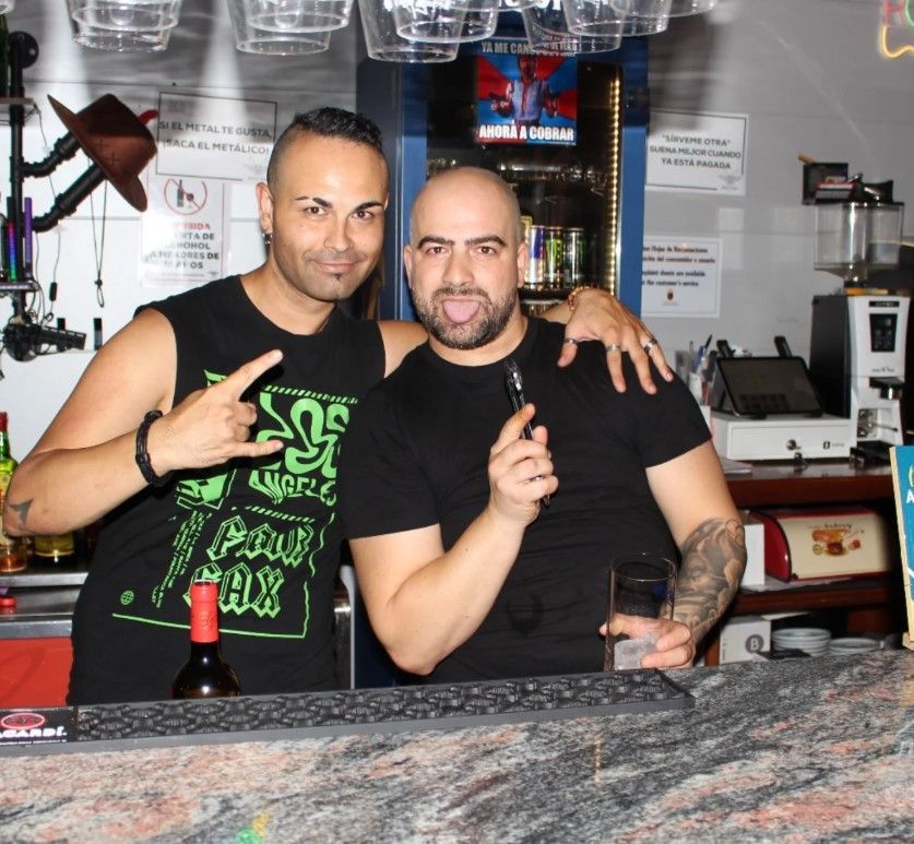 Dos hombres detrás de una barra, uno con un vaporizador en la mano. El otro sonríe con la mano como si fueran cuernos de diablo.
