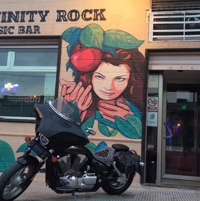 Motocicleta estacionada frente a Infinity Rock Music Bar con un mural de una mujer sosteniendo una manzana.