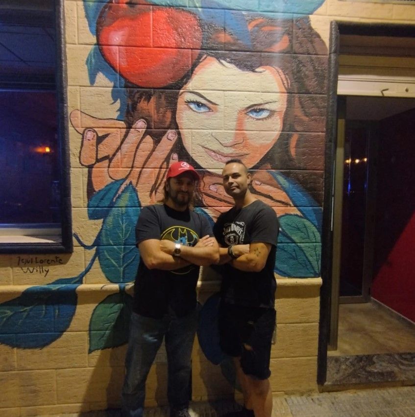 Dos hombres posan con los brazos cruzados frente a un colorido mural que representa a una mujer y una manzana.