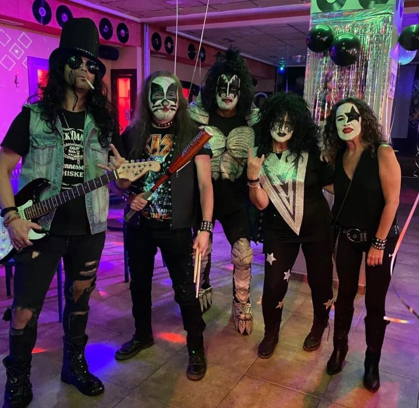 Cinco personas con disfraces de bandas de rock posan en un interior decorado. Uno con sombrero de copa, los demás con la cara pintada.