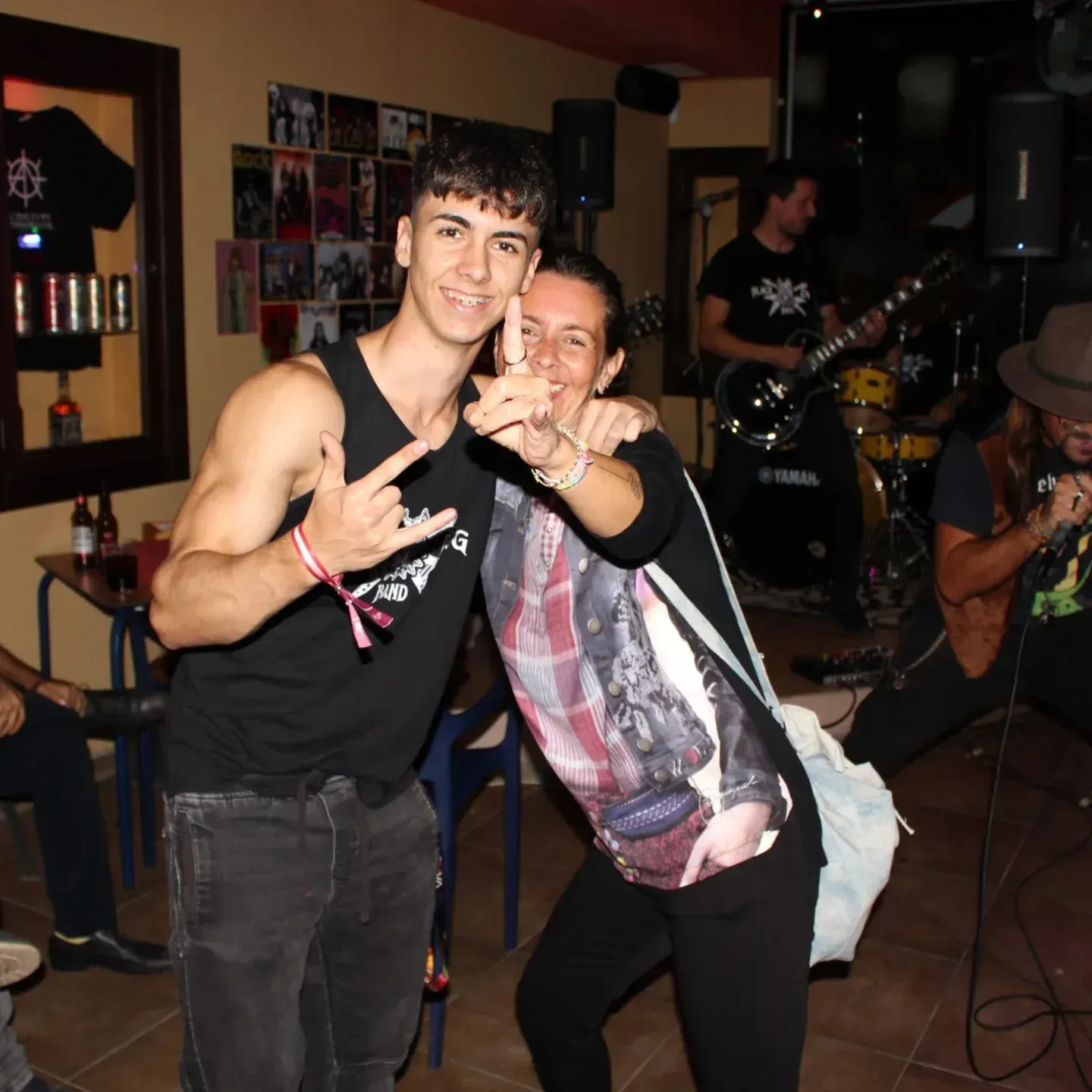 Dos personas posando para una foto en un bar con una banda tocando de fondo, haciendo gestos con las manos.