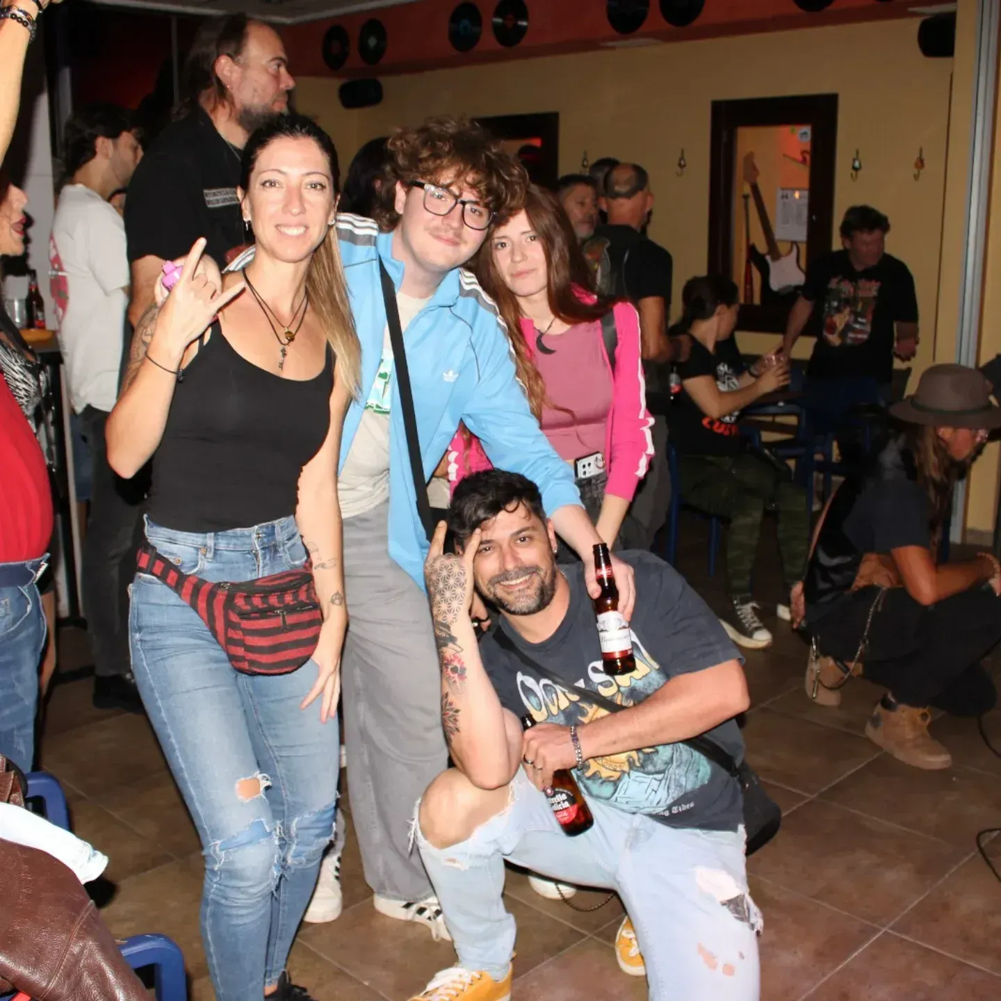 Grupo de personas posando para una foto en un bar; mujer con camiseta negra y hombre sosteniendo cerveza.