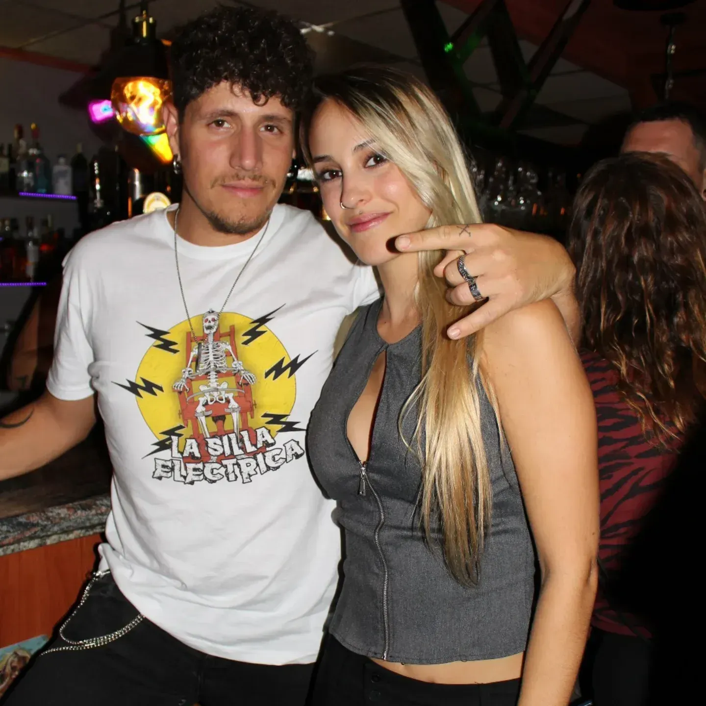 Hombre con camiseta blanca y mujer con chaleco gris posando juntos en un bar.