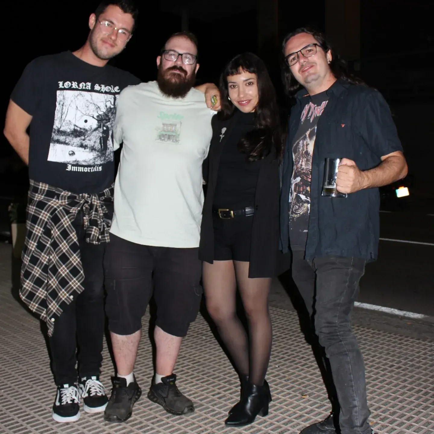Cuatro personas posando al aire libre de noche. El grupo incluye dos hombres y una mujer. Un hombre tiene barba; todos visten ropa oscura.