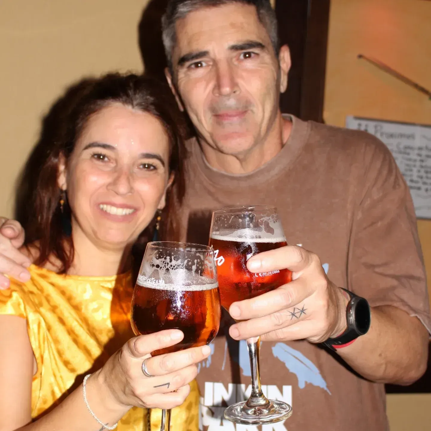Pareja brindando con vasos de cerveza. La mujer de amarillo y el hombre de marrón, ambos sonriendo.