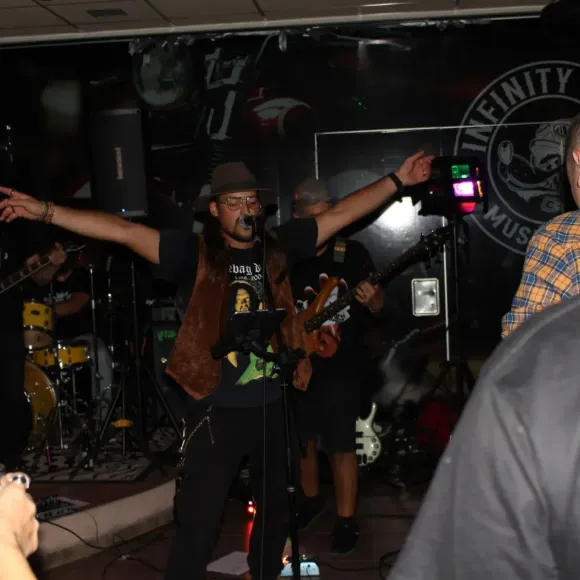 Banda tocando en el escenario: El cantante con los brazos extendidos, con sombrero y chaleco. Otros músicos con instrumentos. El lugar está oscuro.