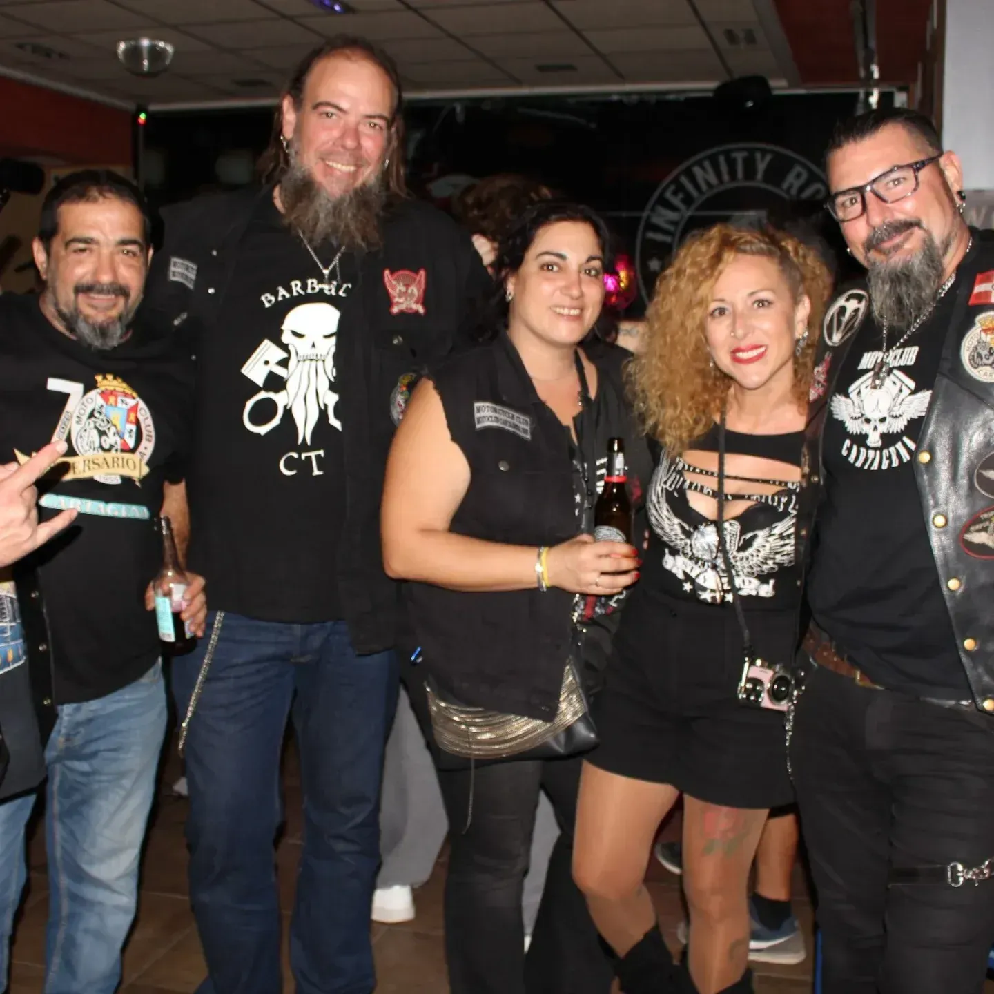 Grupo de personas en un bar, algunas con chalecos con parches. Sonrientes, con bebidas en la mano. El cartel de 