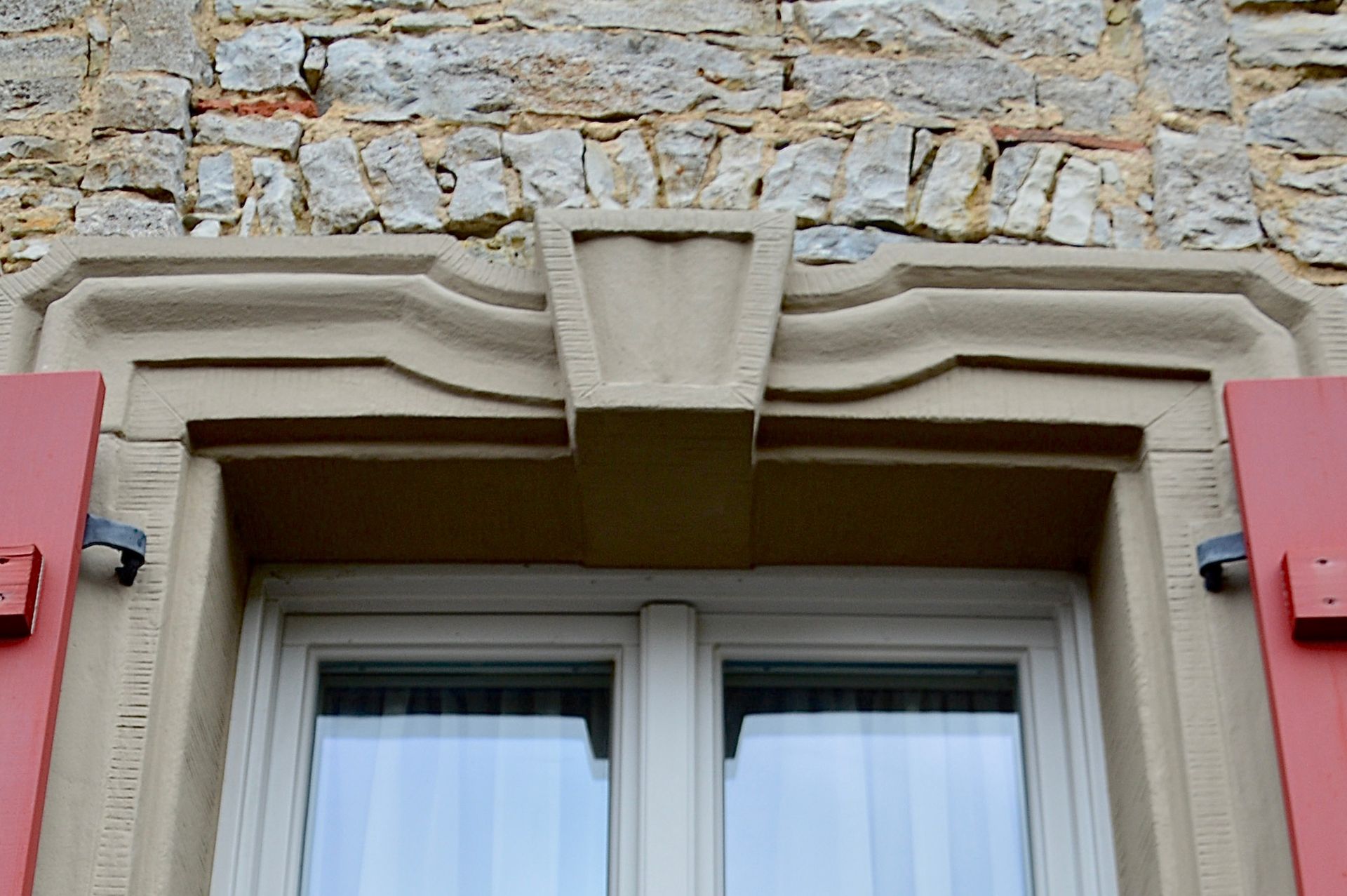 Fenster mit verziertem hellbraunem Steinrahmen, roten Fensterläden, vor dem Hintergrund einer Steinmauer.