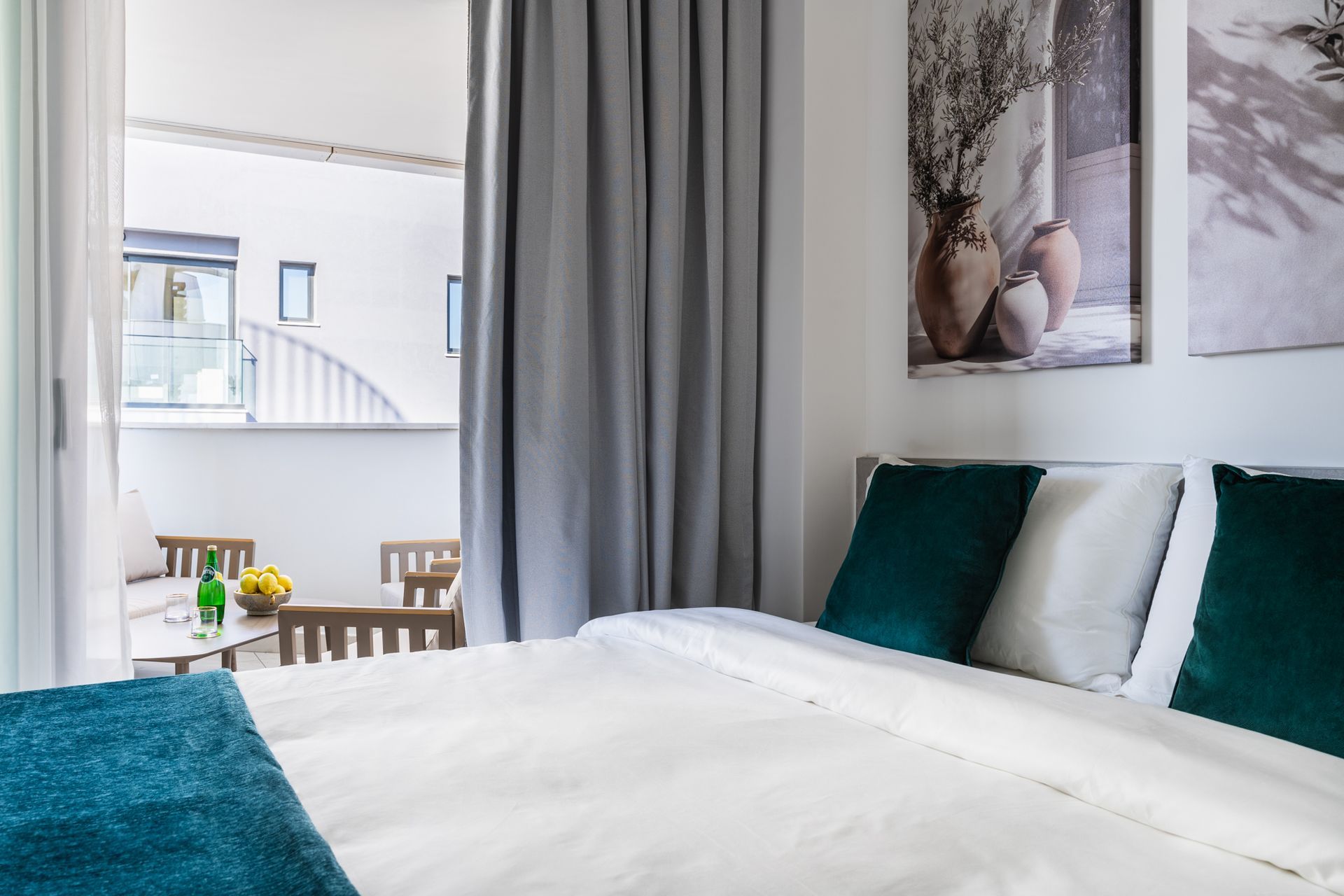 Bedroom with bed, pillows, and balcony access.  Gray curtains, artwork, and outdoor table with fruit.