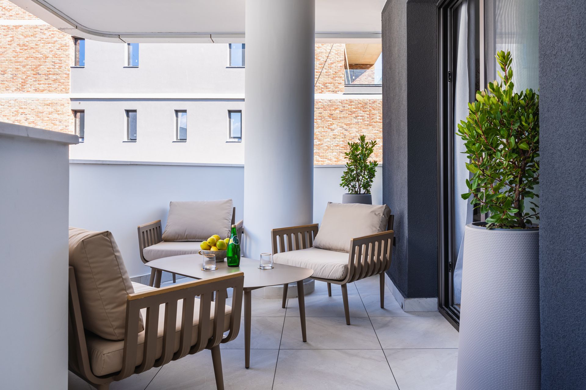 Balcony with seating: two chairs, a small table, a pillar, and potted plants. Building view in background.