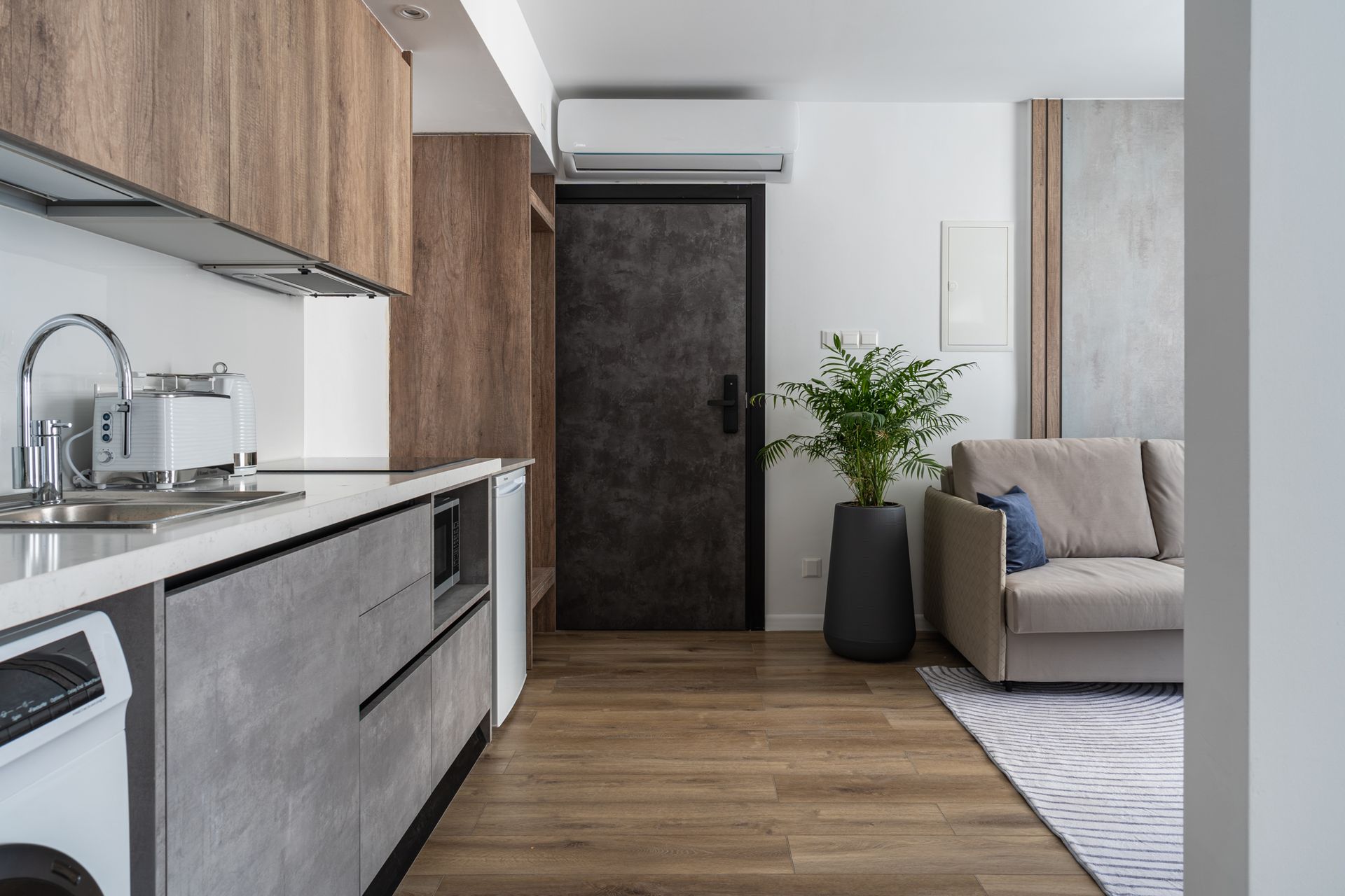Modern kitchen with wood and gray cabinets, and a living area with a sofa, potted plant, and door.