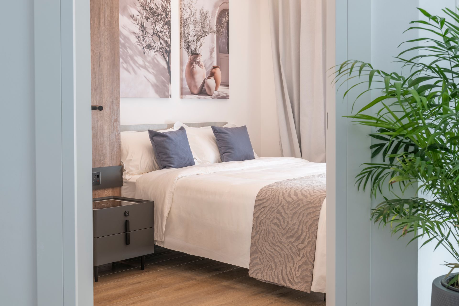 Bedroom interior with a bed, artwork, and a potted plant seen through a doorway.