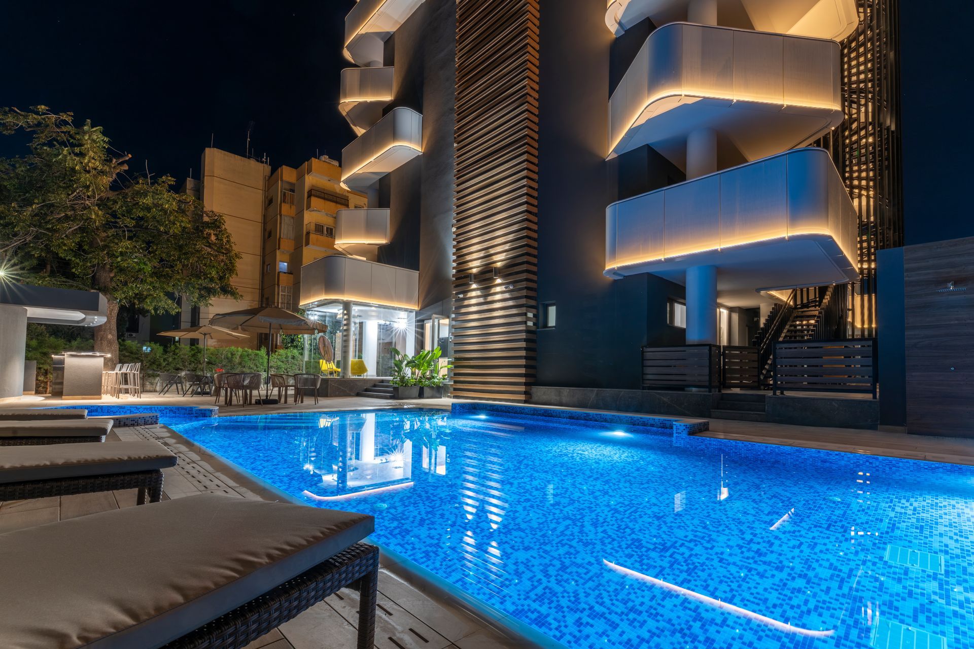 Nighttime view of a pool with blue tile, lounge chairs, and a modern building with lit balconies.