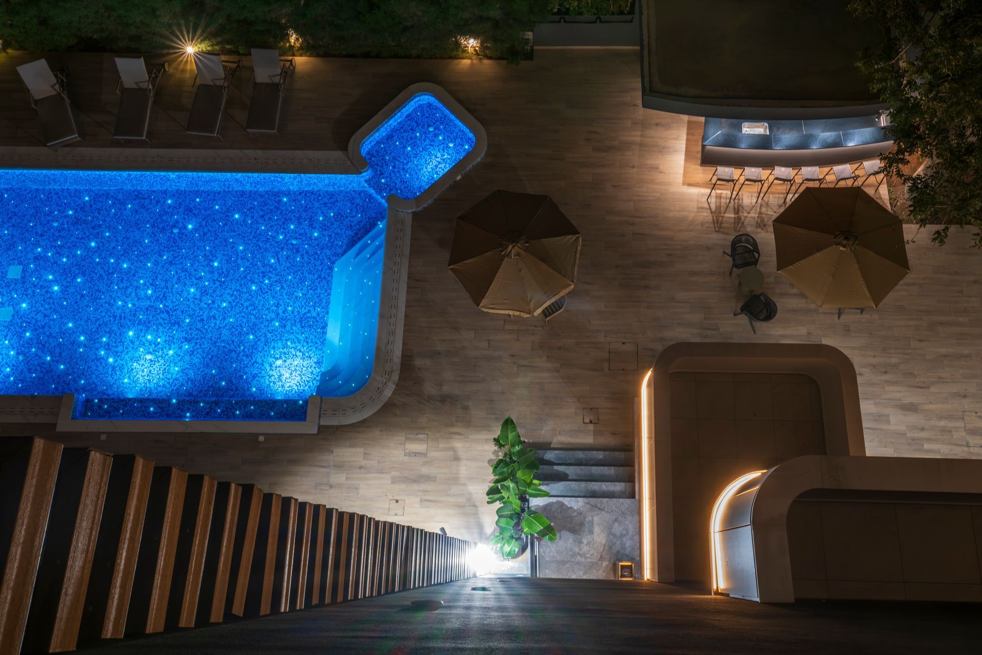 Nighttime aerial view of a blue-lit pool, patio with umbrellas, and a bar area with warm lighting.