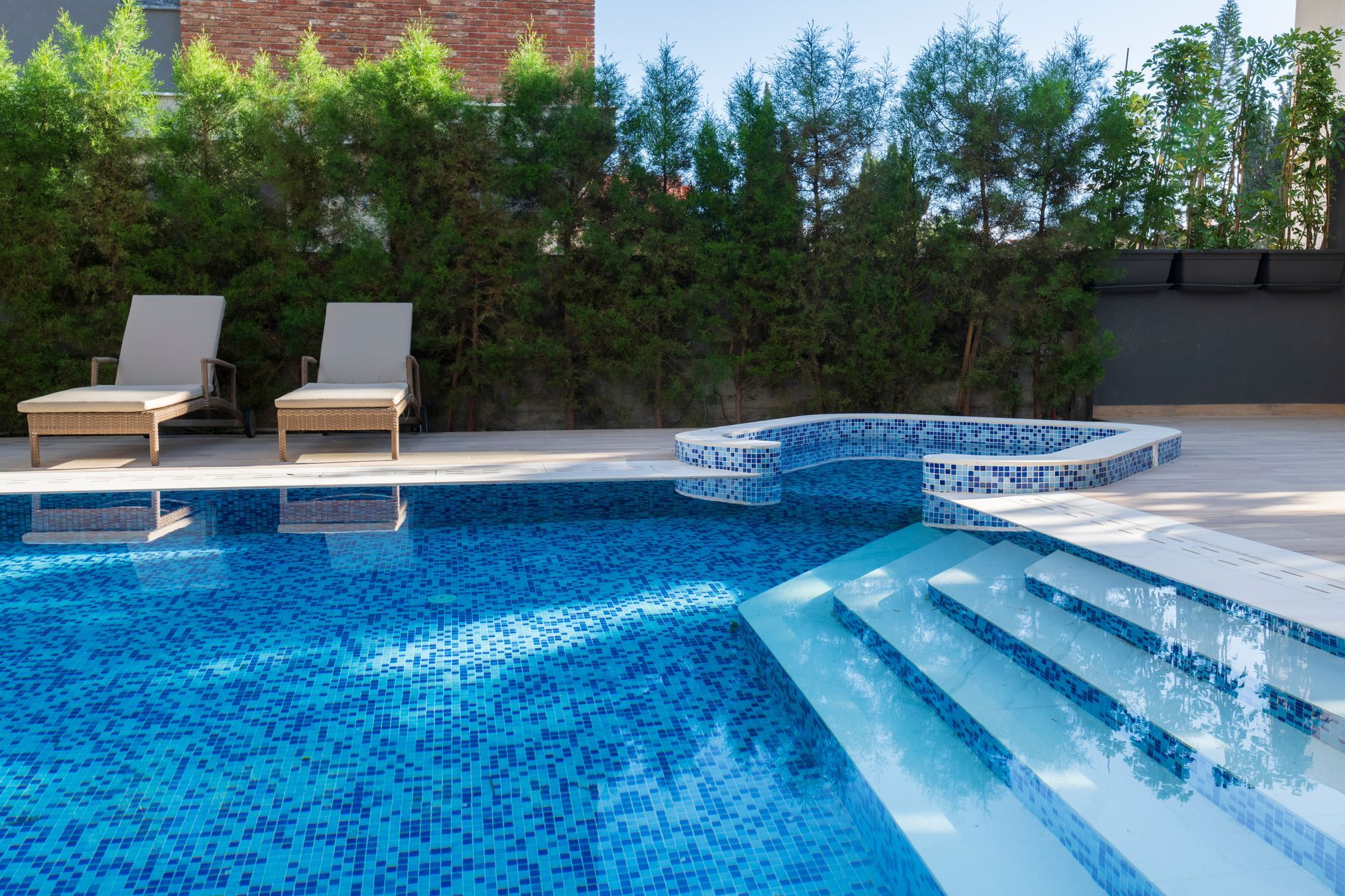Pool with blue mosaic tile, steps, and a jacuzzi. Lounge chairs and greenery in the background.