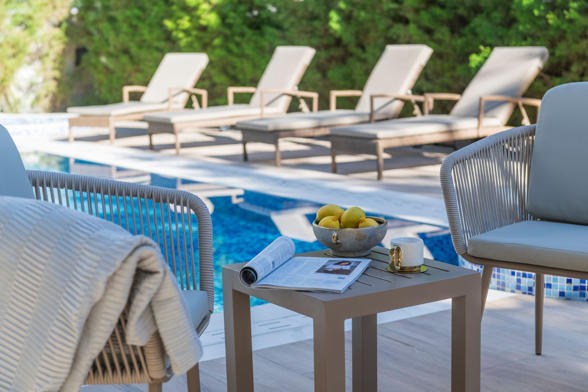 Poolside lounge area with four chaise lounges and two chairs with a small table, a bowl of lemons, and a magazine.