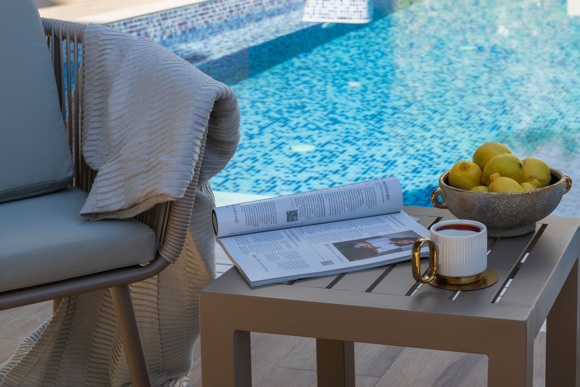 A chair with a blanket, table with a magazine, a cup, and a bowl of lemons beside a pool.