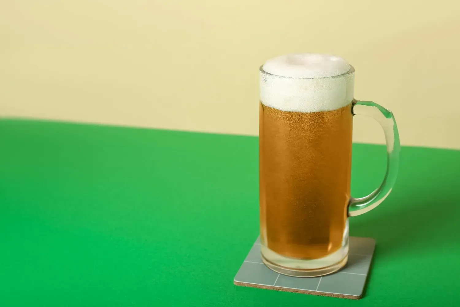 Vaso alto de cerveza con espuma sobre un posavasos cuadrado sobre un fondo verde de mesa.
