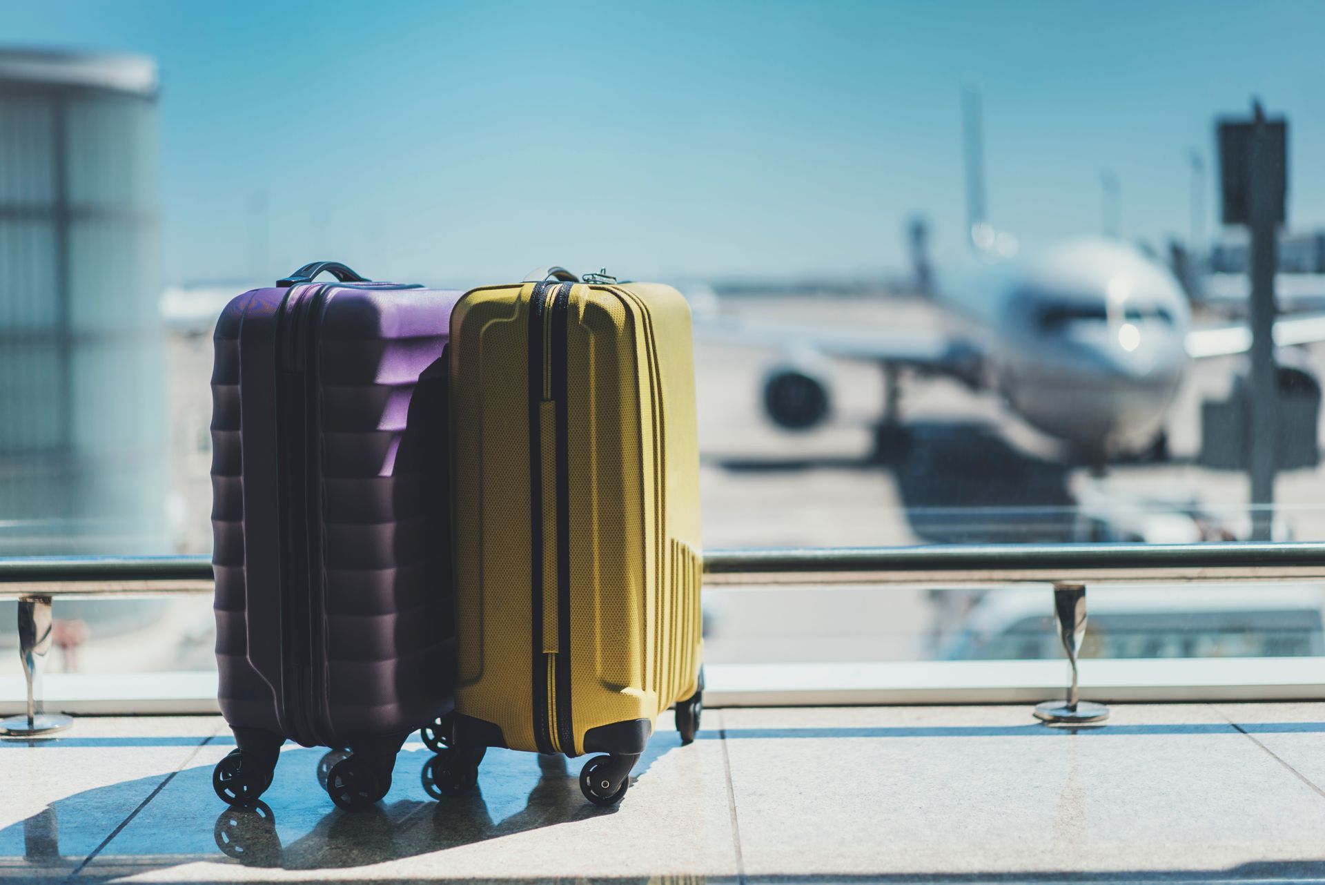 Une valise violette et une valise jaune sont côte à côte sur le sol d'un aéroport, devant une fenêtre donnant sur un avion stationné.