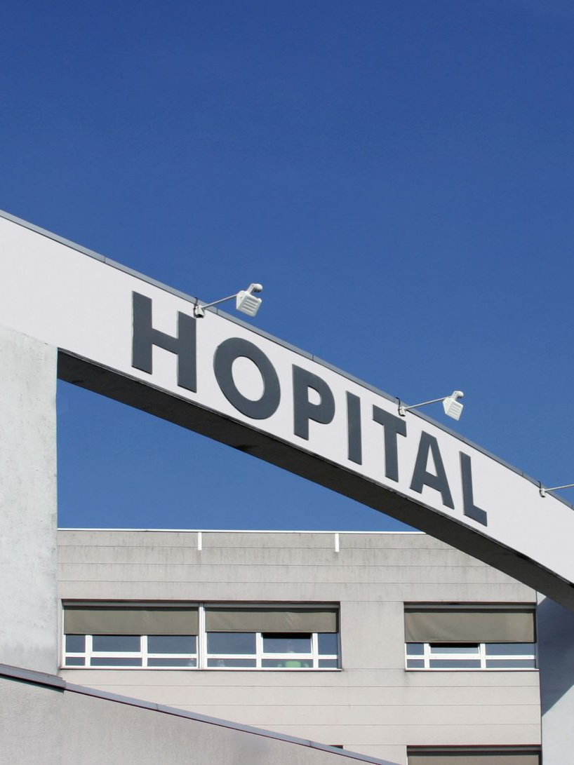 Une grande enseigne blanche inclinée affichant le mot HÔPITAL en lettres noires grasses sur fond de ciel bleu clair et d'une aile d'hôpital.