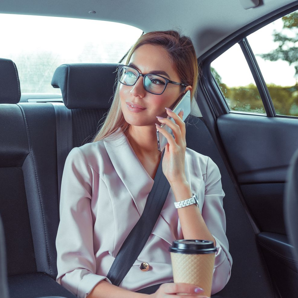 Une personne vêtue d'un blazer rose clair et portant des lunettes parle au téléphone tout en tenant un gobelet de café sur la banquette arrière d'une voiture.