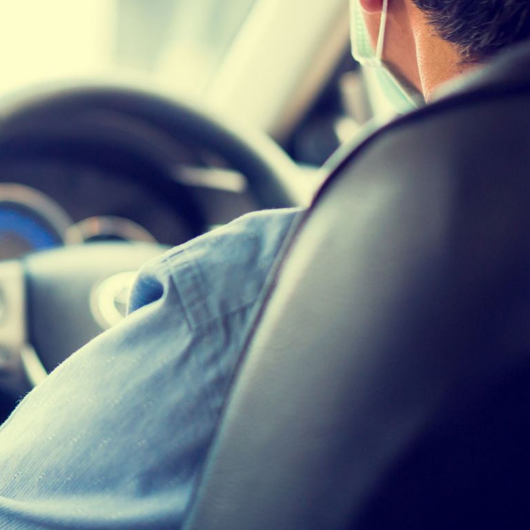 Une personne portant une chemise bleue et un masque est assise sur le siège conducteur d'une voiture, vue de derrière.