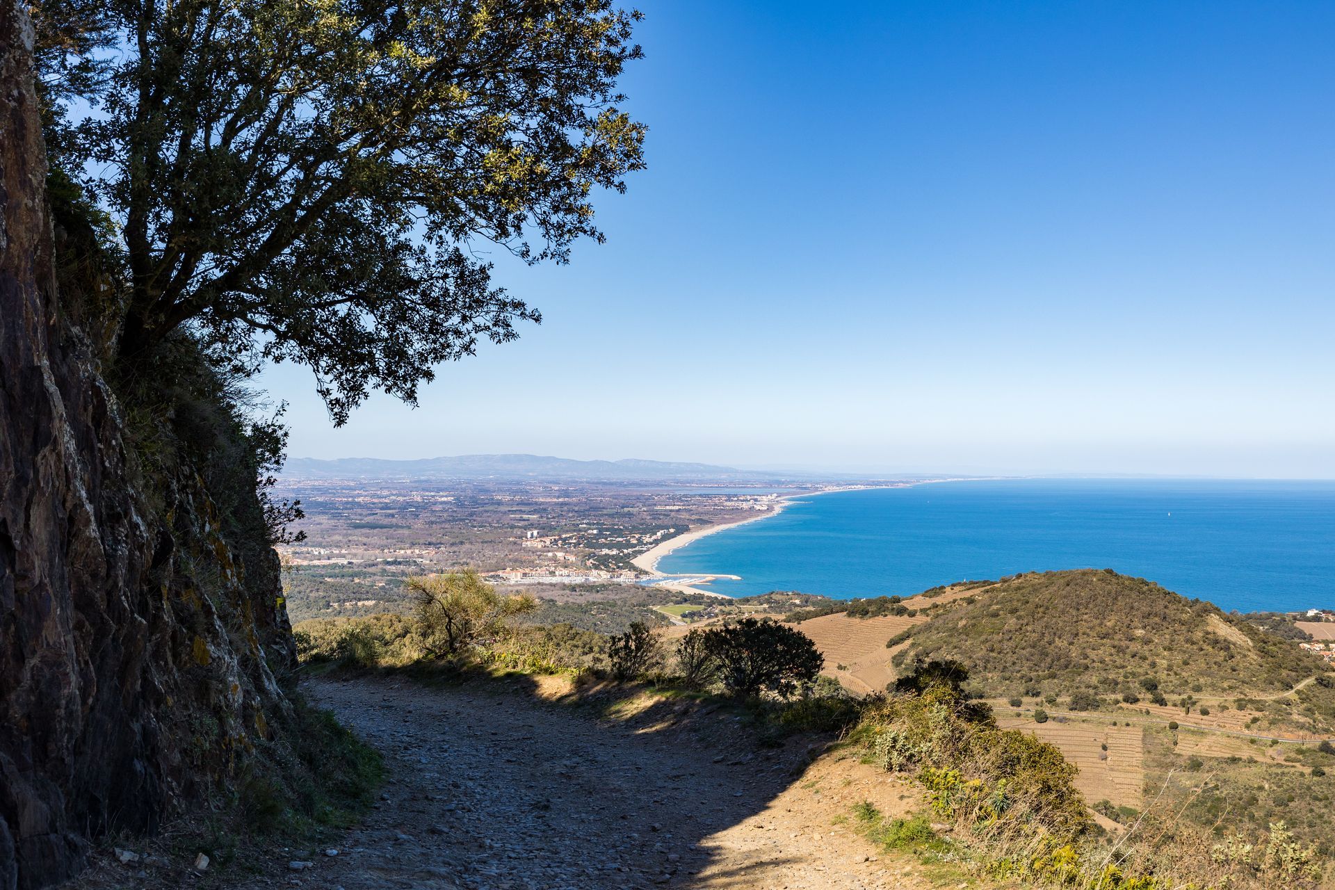 Vue depuis un chemin de terre à flanc de colline surplombant une ville côtière et la mer Méditerranée d'un bleu profond sous un ciel dégagé.