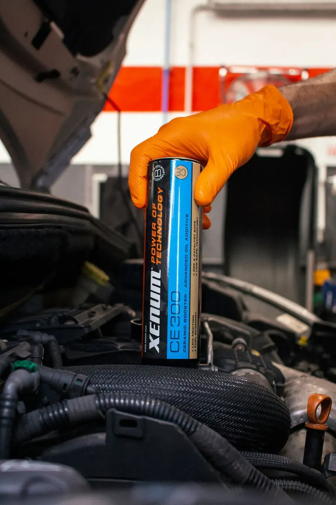Mano enguantada sosteniendo un recipiente cilíndrico sobre un motor de automóvil; el recipiente es negro y azul, etiquetado como