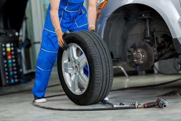 Mecánico con mono azul, sosteniendo un neumático, trabajando en un coche plateado en un taller.