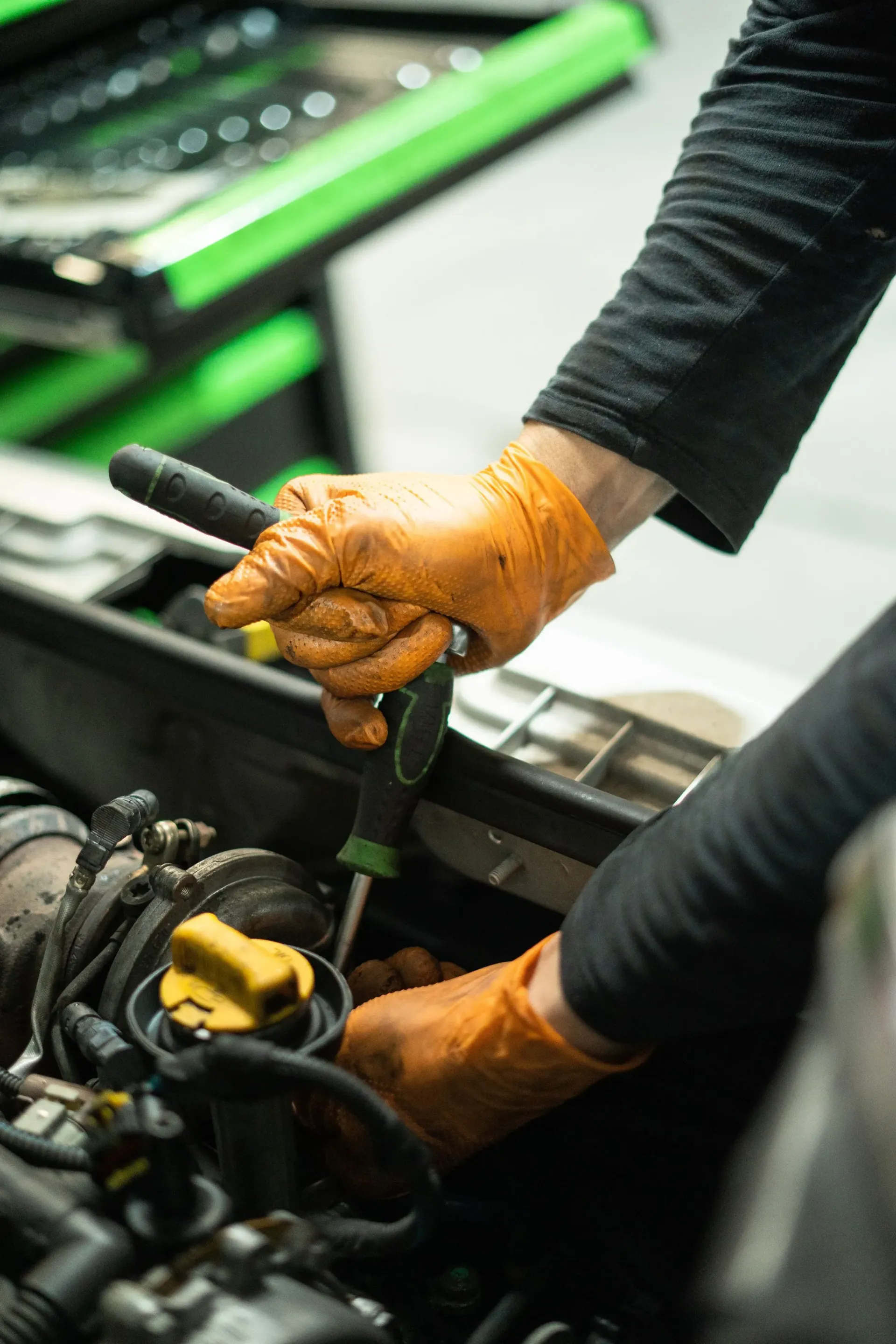 Manos con guantes naranjas usando un destornillador en el motor de un coche. Caja de herramientas verde al fondo.