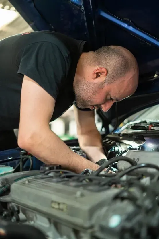 Hombre trabajando en el motor de un coche con el capó abierto, vestido con una camisa negra, en el interior de un edificio.