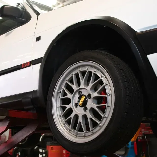 Coche blanco con llanta plateada elevado sobre un elevador, con guardabarros negros y pinza de freno roja visible.
