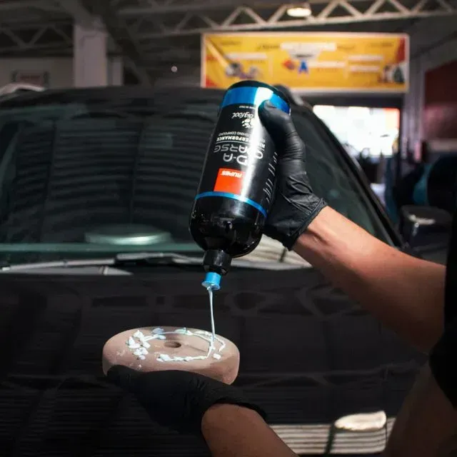 Persona con guantes negros vertiendo líquido de una botella sobre una almohadilla de pulido en el capó de un coche.