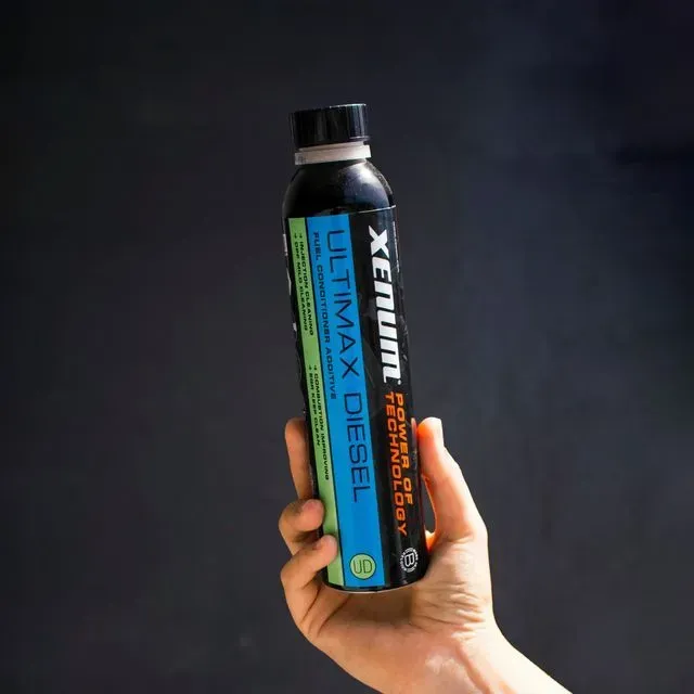 Mano sosteniendo una botella de aditivo para combustible diésel XENUM Ultimax. Botella negra, etiqueta verde y azul.