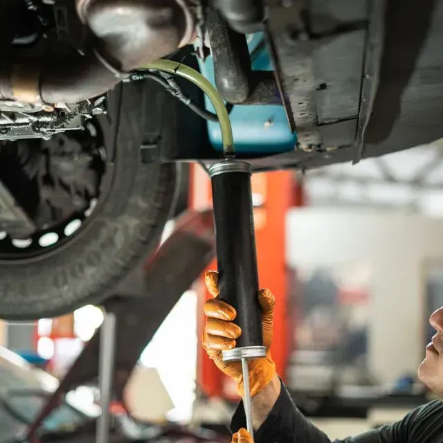 Mecánico extrayendo fluido de un vehículo con una bomba. Manos con guantes naranjas, en un taller.