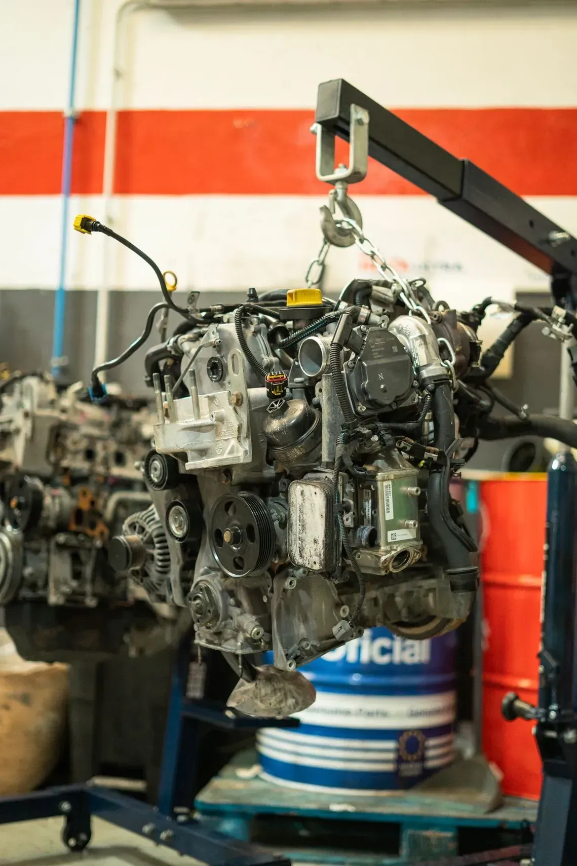 Motor izado con una grúa en un taller de reparación, con otro motor al fondo y un tambor de pintura debajo.