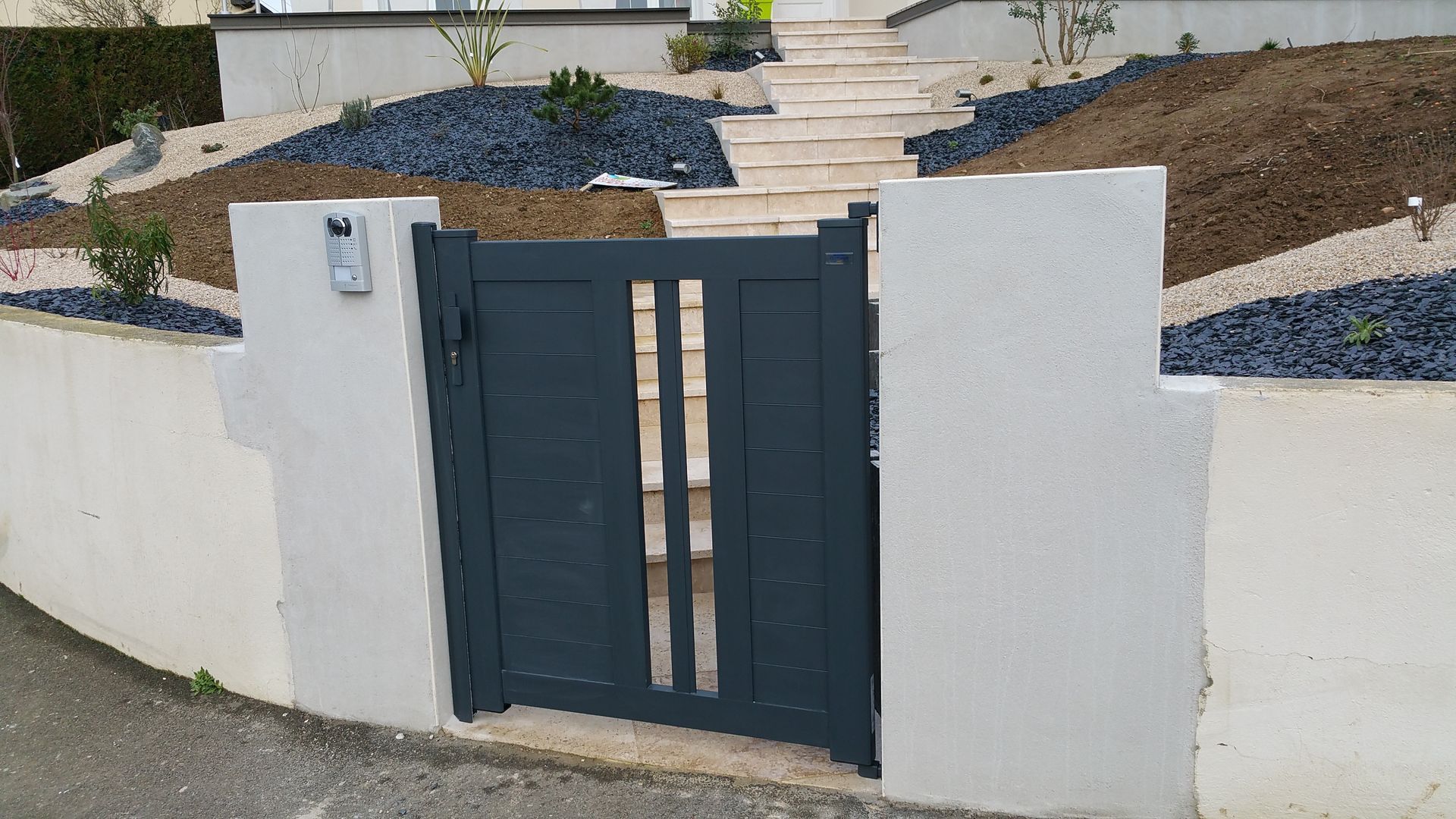 Un portail en métal gris foncé encastré dans un mur blanc, avec des marches menant à une cour paysagée recouverte de paillis de gravier foncé.