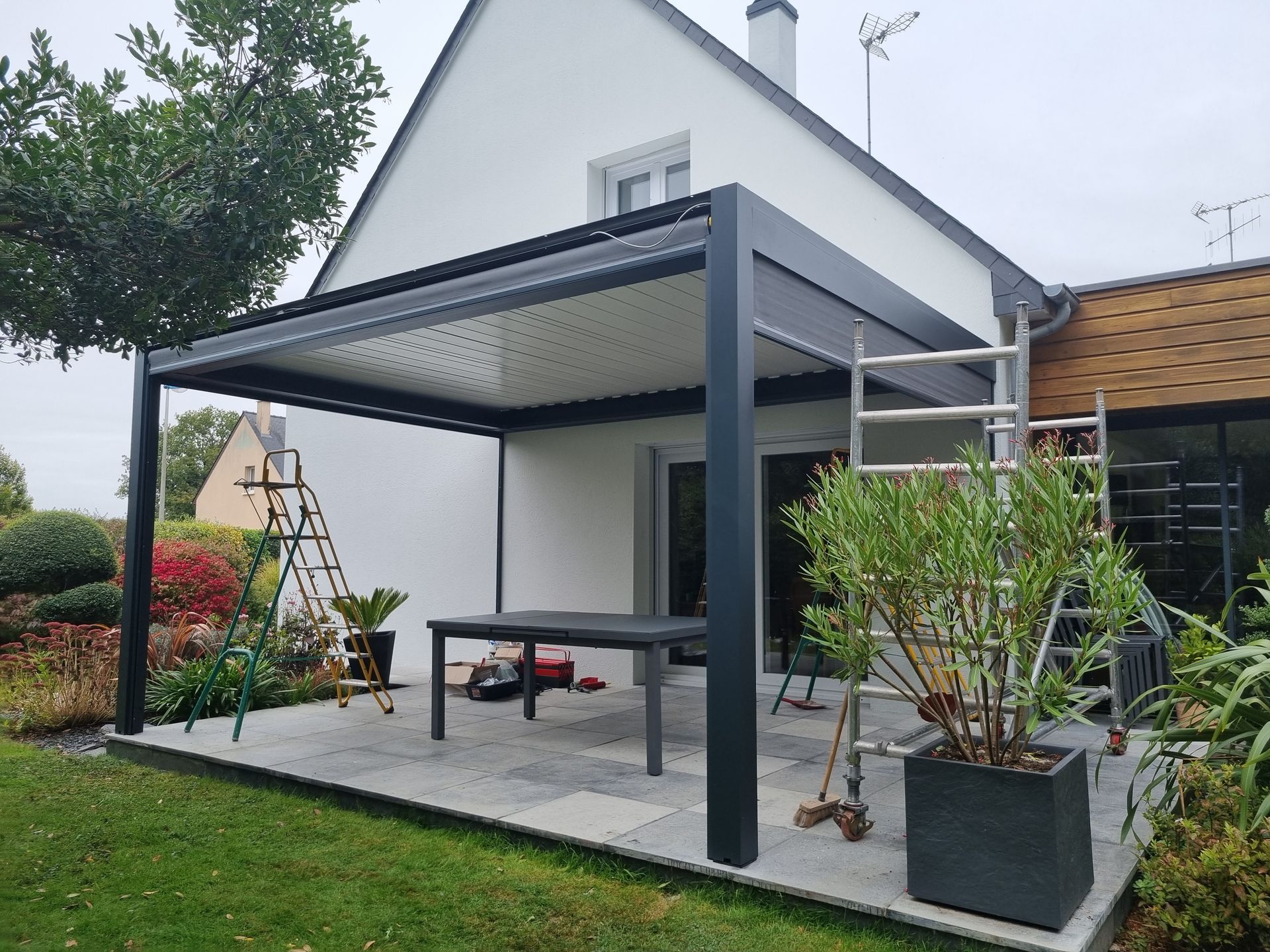 Une pergola moderne gris foncé, avec un toit à claire-voie, couvre un patio devant une maison blanche ; une échelle et des plantes en pot y sont disposées.
