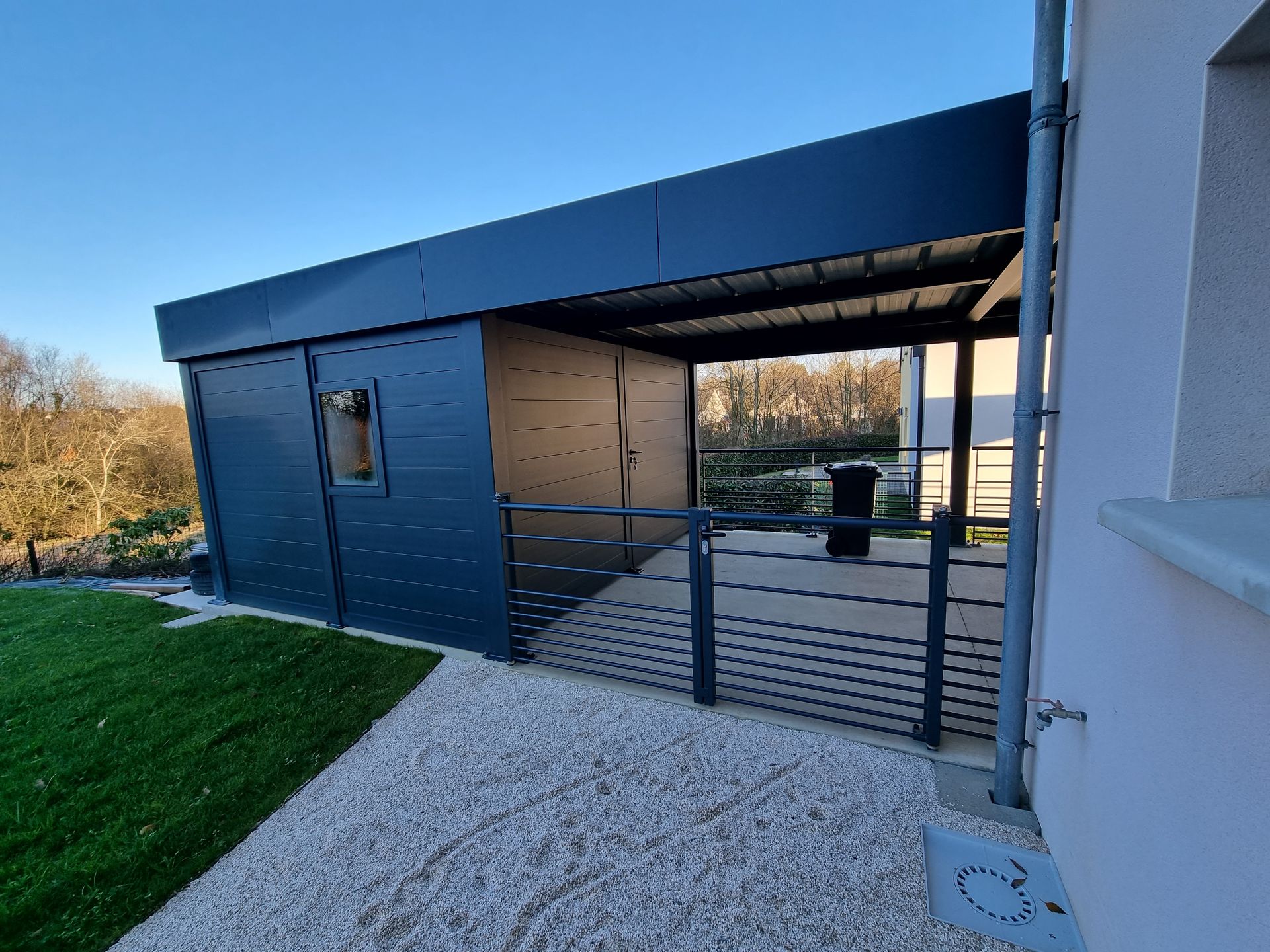 Un abri de voiture ou une structure de patio extérieure moderne, de couleur bleu foncé, avec un toit plat, posée sur du gravier à côté d'un mur de maison.
