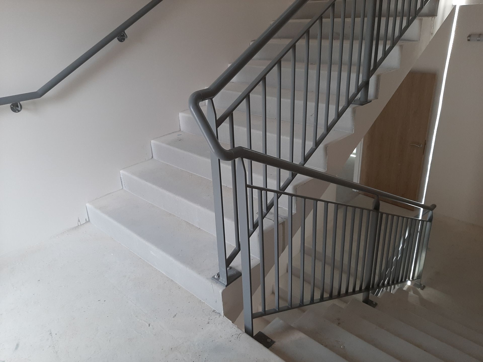 Escalier avec marches en béton clair, murs blancs et rampes en métal foncé dans une cage d'escalier moderne.