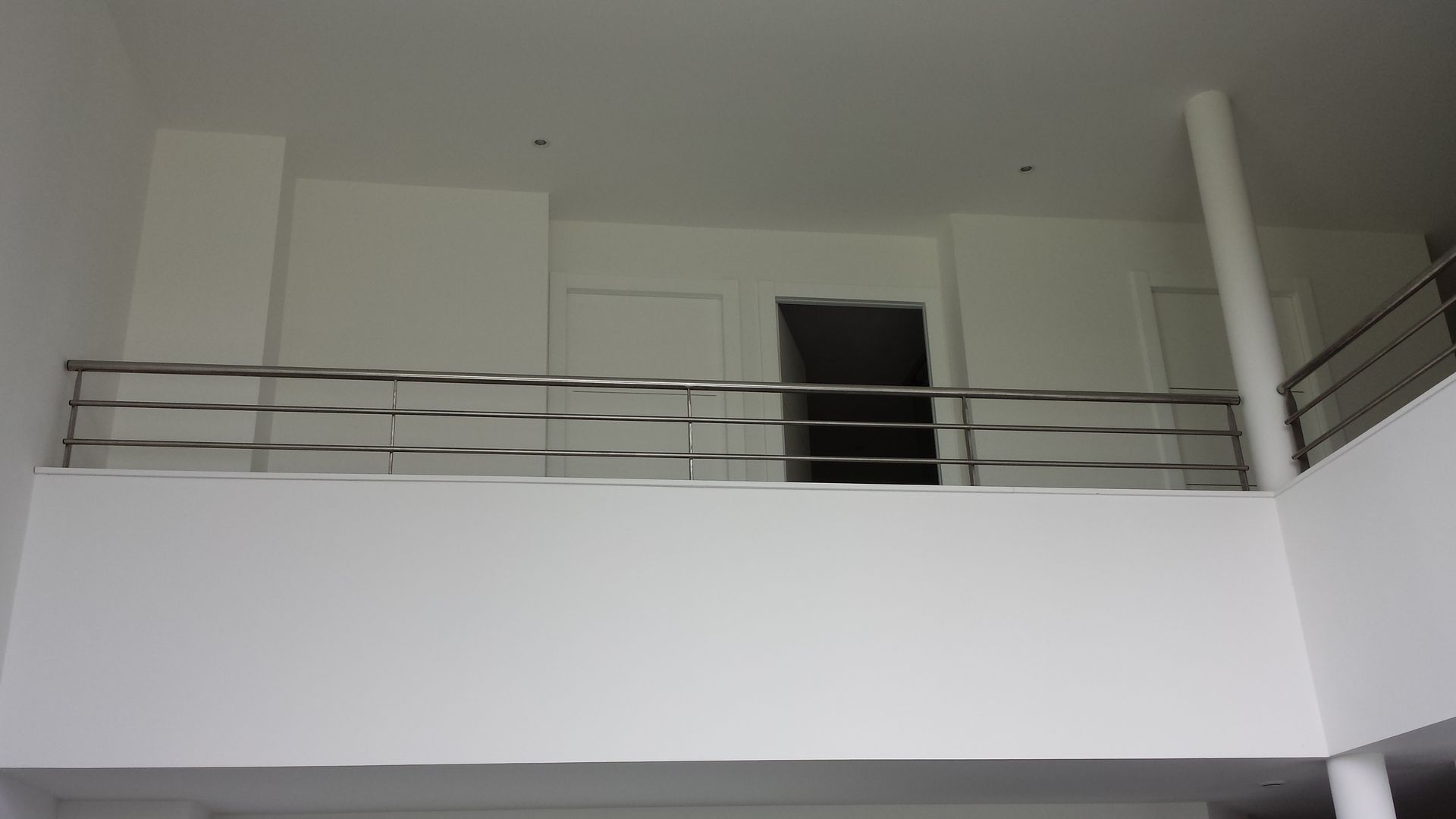 Un balcon intérieur blanc avec une rambarde métallique horizontale, donnant sur une petite porte à l'étage supérieur.