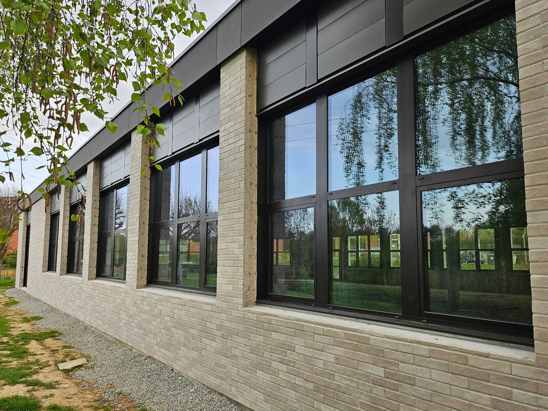 L'extérieur d'un bâtiment moderne se caractérise par des murs en briques claires, de grandes fenêtres à cadre sombre et une toiture en métal foncé.